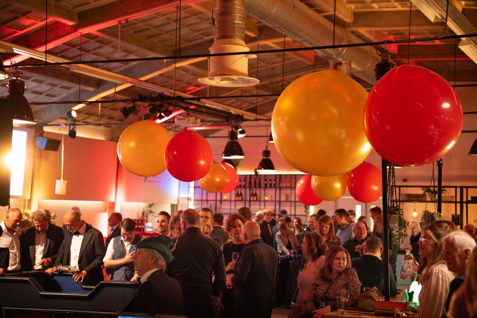 Zaaloverzicht met rode en gele ballonnen die boven de gasten hangen, links een speeltafel en rechts de bar met flessen in de schappen