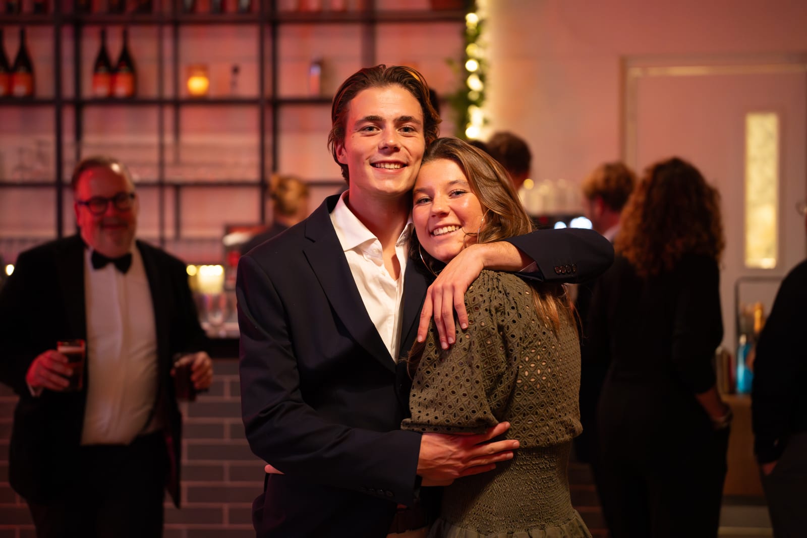 Jong stel poseert lachend met een arm om elkaar heen voor de bar, met champagneflessen in de verlichte schappen erachter