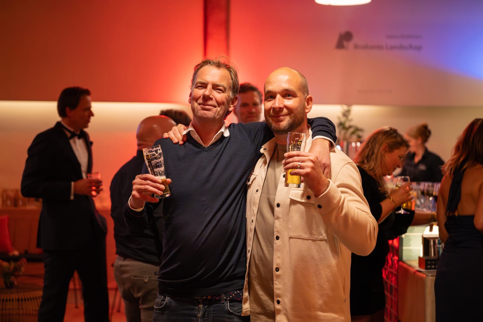 Twee mannen poseren met een arm om elkaar heen en een glas bier in de hand, in de rood verlichte ontvangstruimte