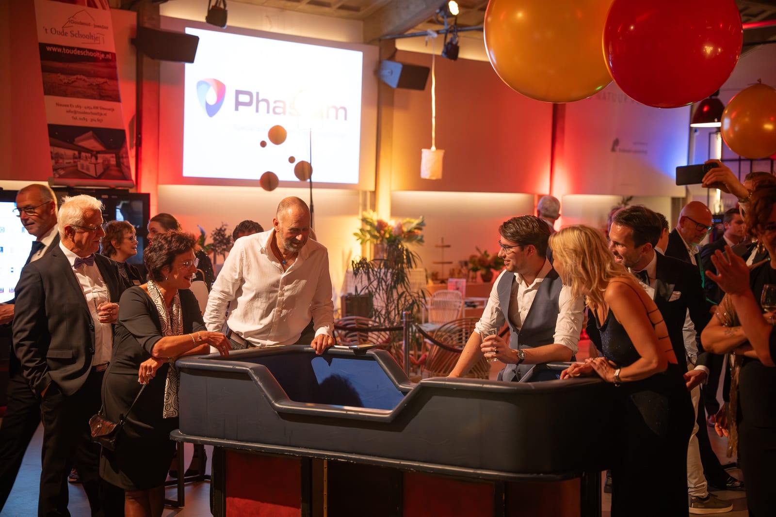Gasten staan rond de speeltafel terwijl op het grote scherm erachter een sponsorlogo wordt geprojecteerd, met ballonnen boven de zaal
