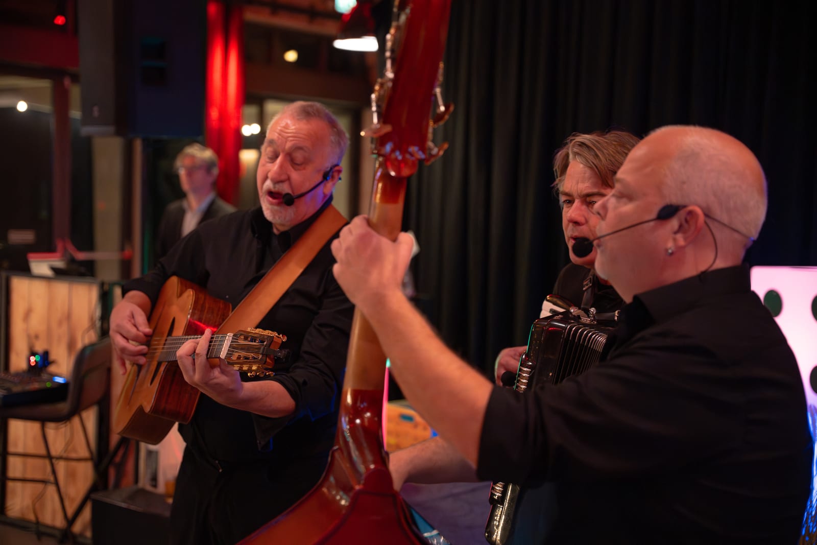 De liveband in actie met gitarist, contrabassist en accordeonist, gezien vanaf de zijkant van het podium in rood en blauw licht