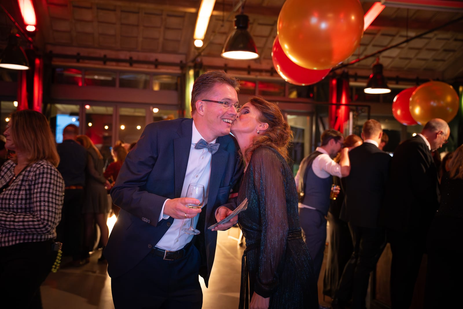 Vrouw geeft een man in smoking een kus op de wang terwijl hij een glas champagne vasthoudt, met rode en oranje ballonnen boven de gasten