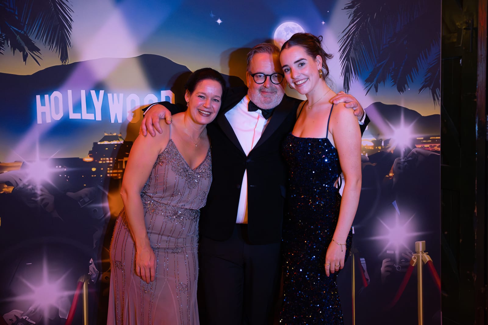 Twee vrouwen in glinsterende avondjurken en een man in smoking poseren voor de fotowand met het Hollywood-decor
