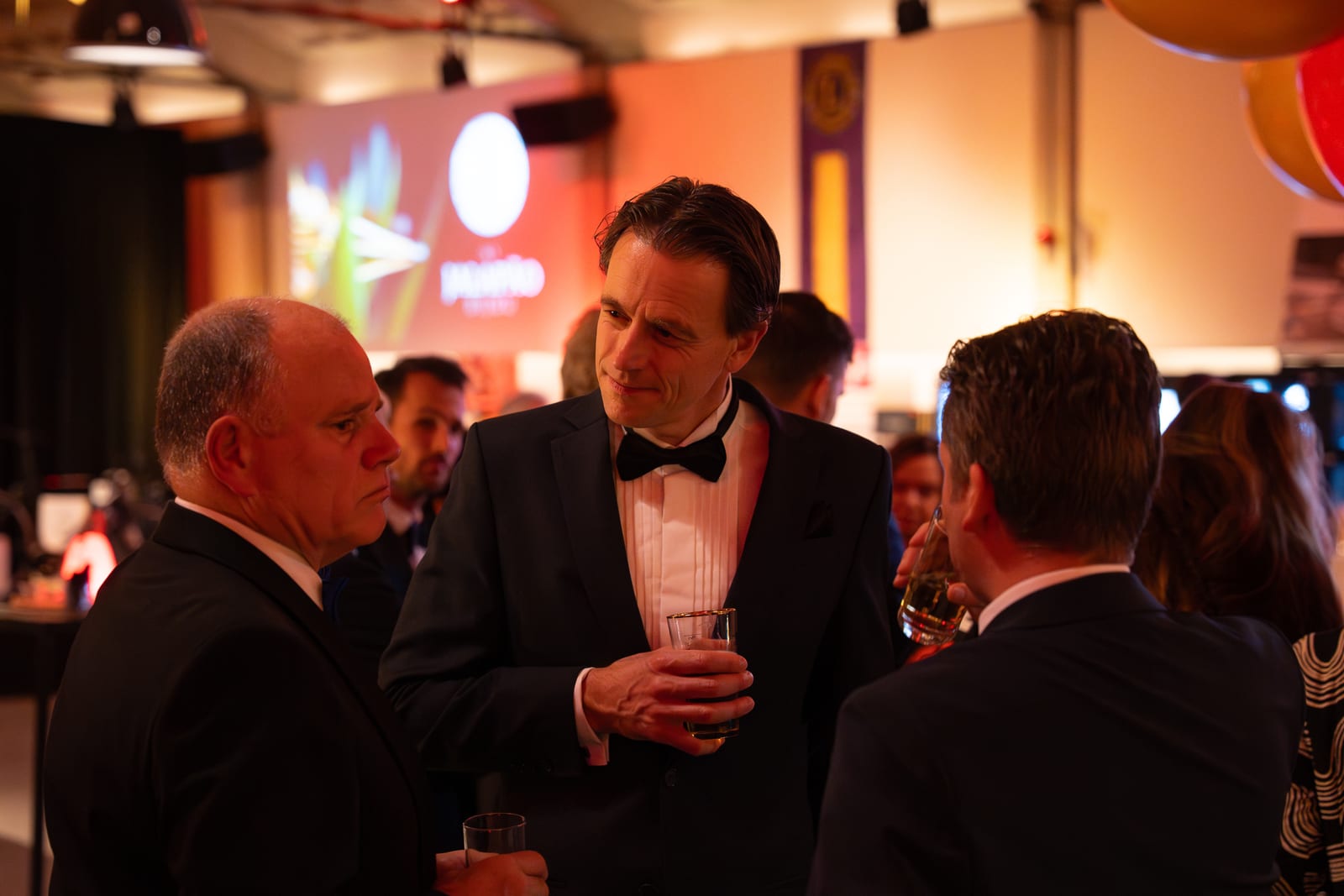 Drie mannen in smoking met vlinderdas staan met een glas in gesprek, met een verlicht projectiescherm en het Lions-vaandel achter hen