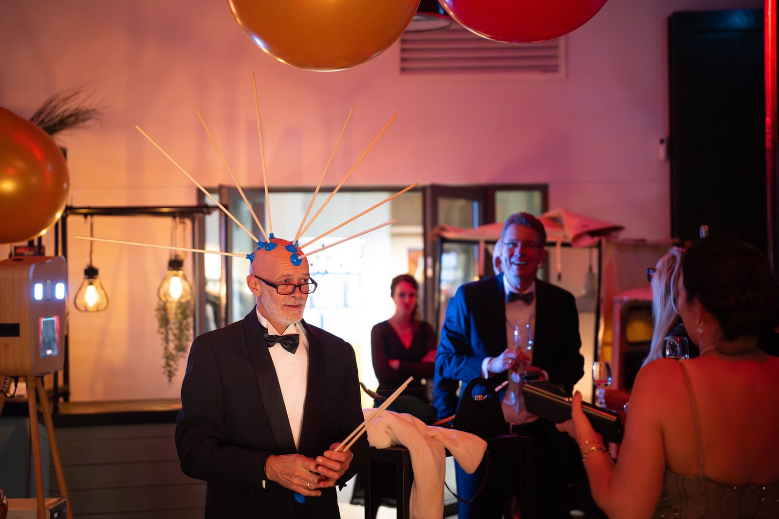 Man in smoking draagt lachend een opvallende hoofdtooi met uitstekende sprieten en houdt sterretjes vast, met ballonnen boven hem