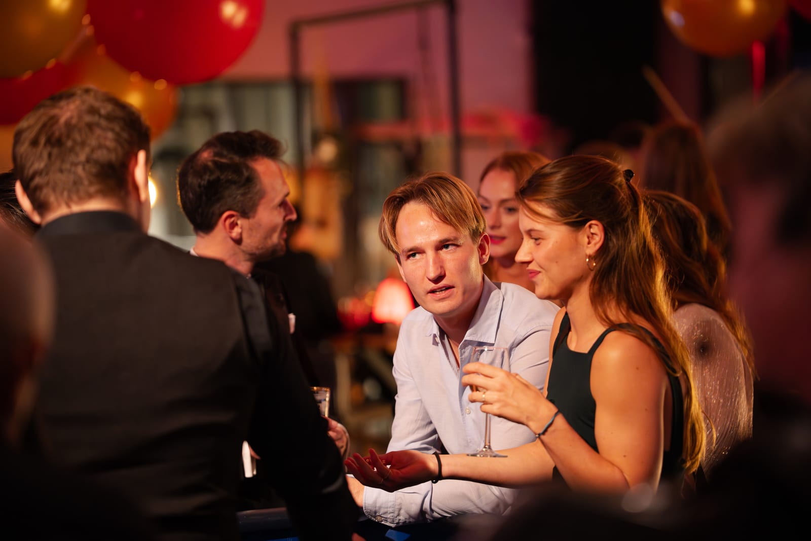 Een man en vrouw zitten dicht bij elkaar met een glas champagne, omringd door pratende gasten en rode ballonnen