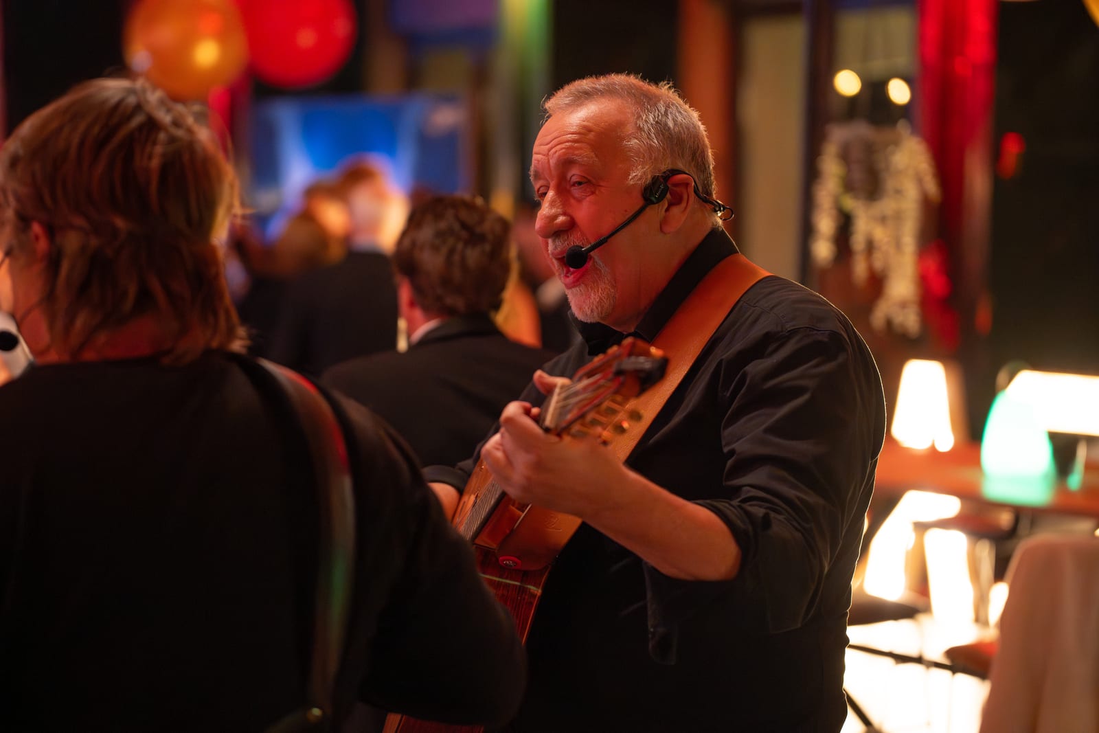 Zingende gitarist met headsetmicrofoon speelt tussen de gasten in de zaal, met ballonnen en schemerlampen op de achtergrond