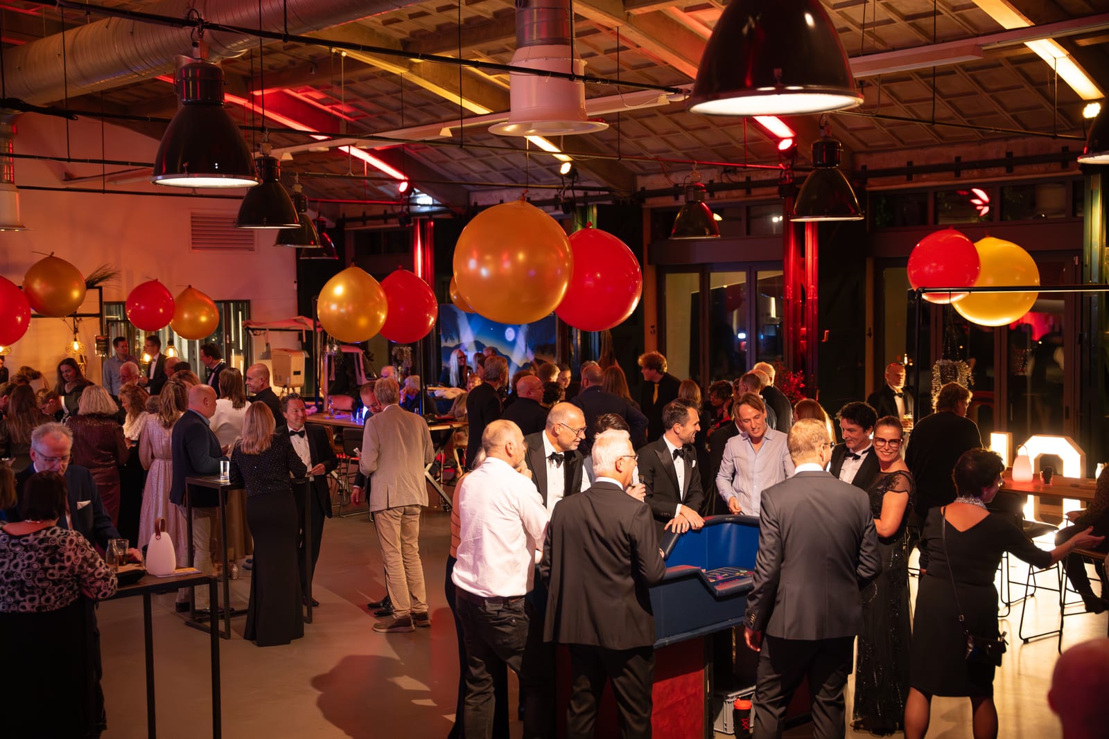 Gasten verzameld rond de blauwe blackjacktafel midden in de zaal, met ballonnen en industriële hanglampen aan het plafond