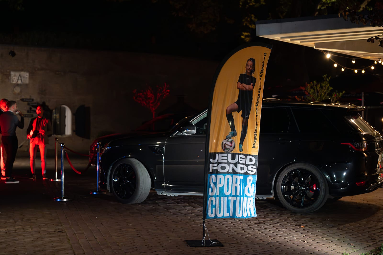 Beachflag van Jeugdfonds Sport & Cultuur staat 's avonds buiten bij de ingang, naast een zwarte terreinwagen en het rode koord