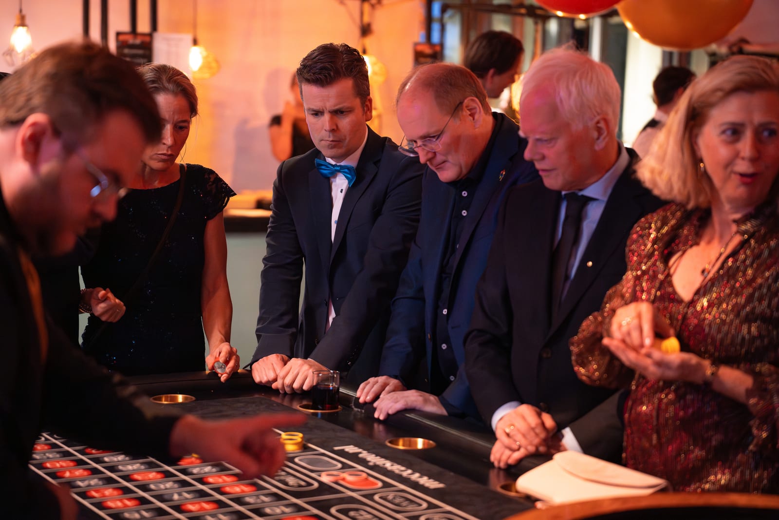 Gasten in avondkleding staan geconcentreerd rond de roulettetafel terwijl de croupier op de voorgrond het spel leidt