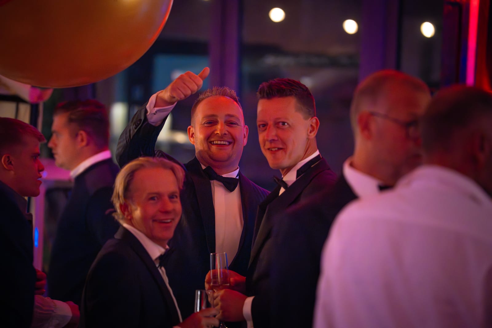 Drie mannen in smoking lachen naar de camera, een van hen steekt zijn duim op, in paars en rood feestlicht