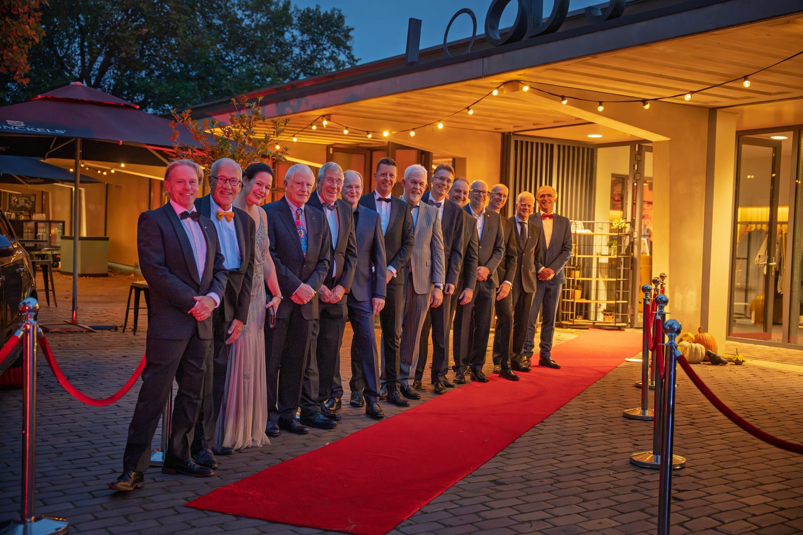 Vijftien leden van de organisatie staan in een rij langs de rode loper voor de entree, onder een sliert lampjes in de schemering