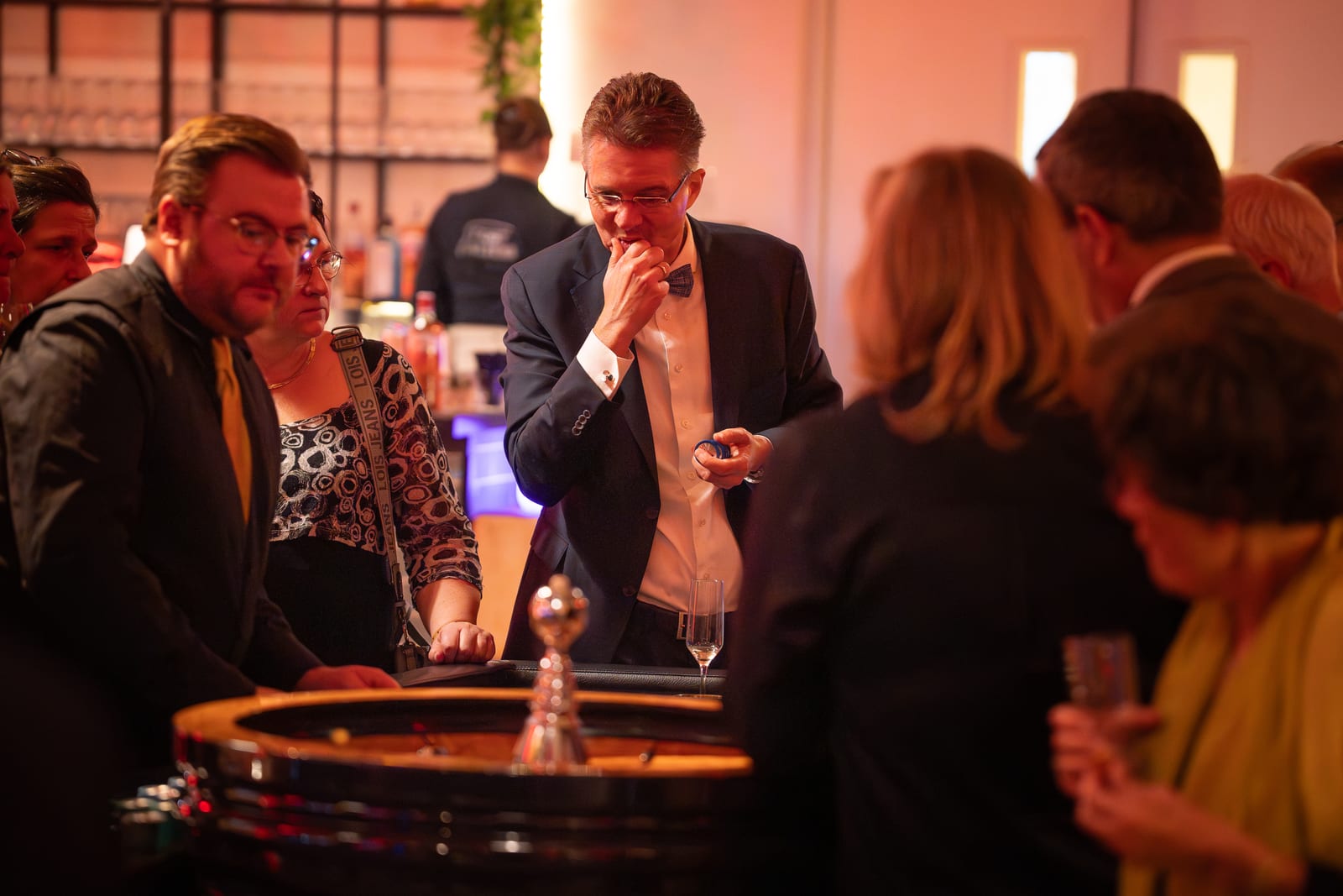 Man met vlinderdas bekijkt bedachtzaam zijn fiche aan de roulettetafel, omringd door medespelers in het warme zaallicht