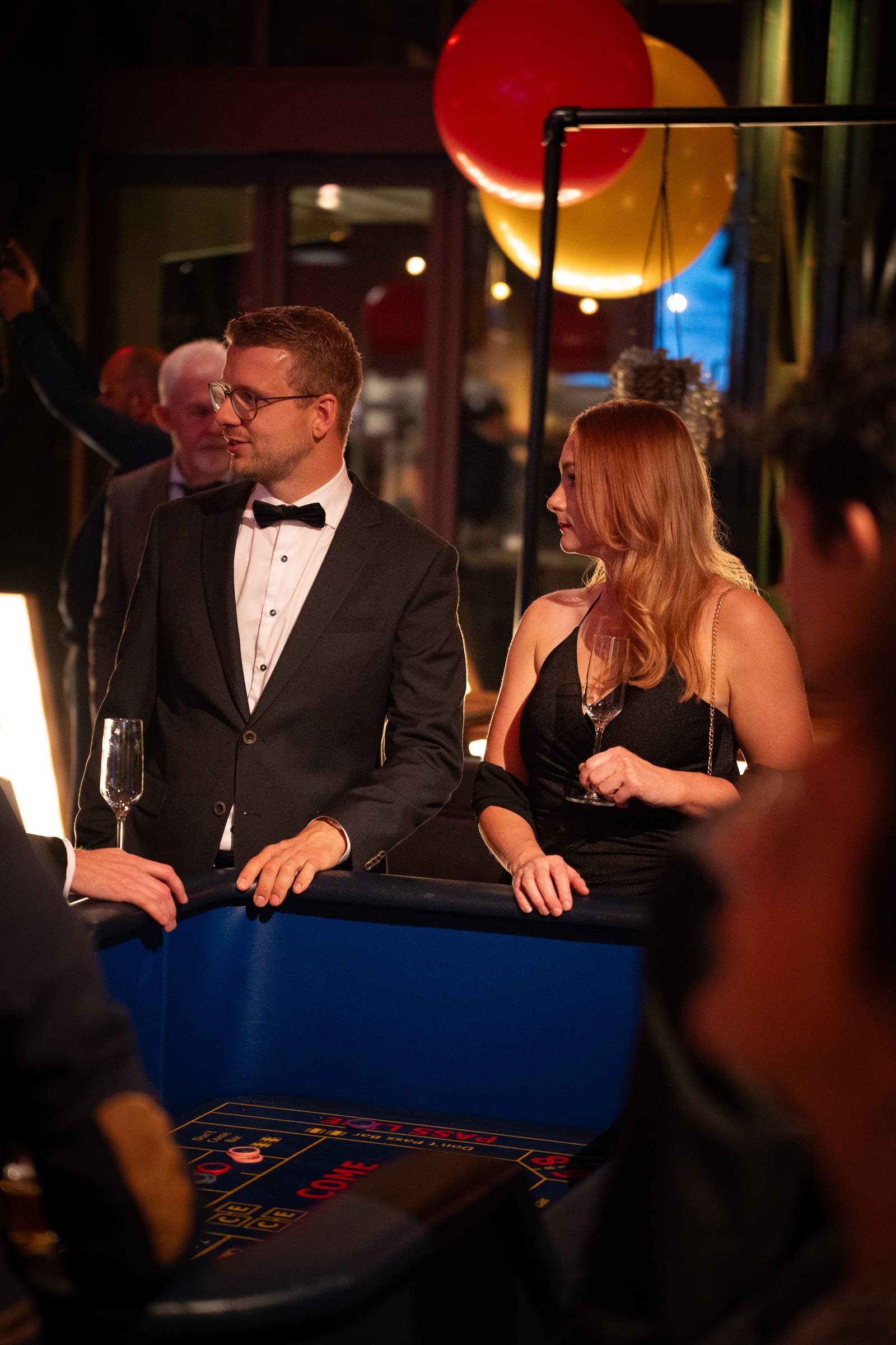 Staand portret van een man in smoking met vlinderdas en een vrouw in zwarte jurk aan de blauwe craps-tafel, met rode en gele ballonnen boven hen