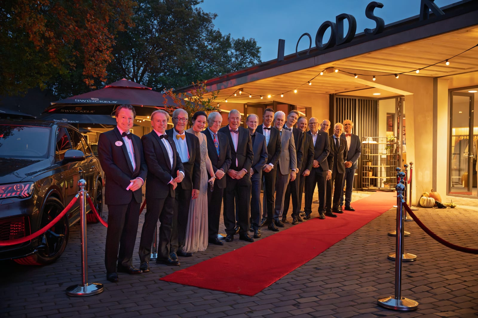 Wijde opname van de organisatie langs de rode loper voor de entree in de schemering, met een auto en rode koorden links in beeld