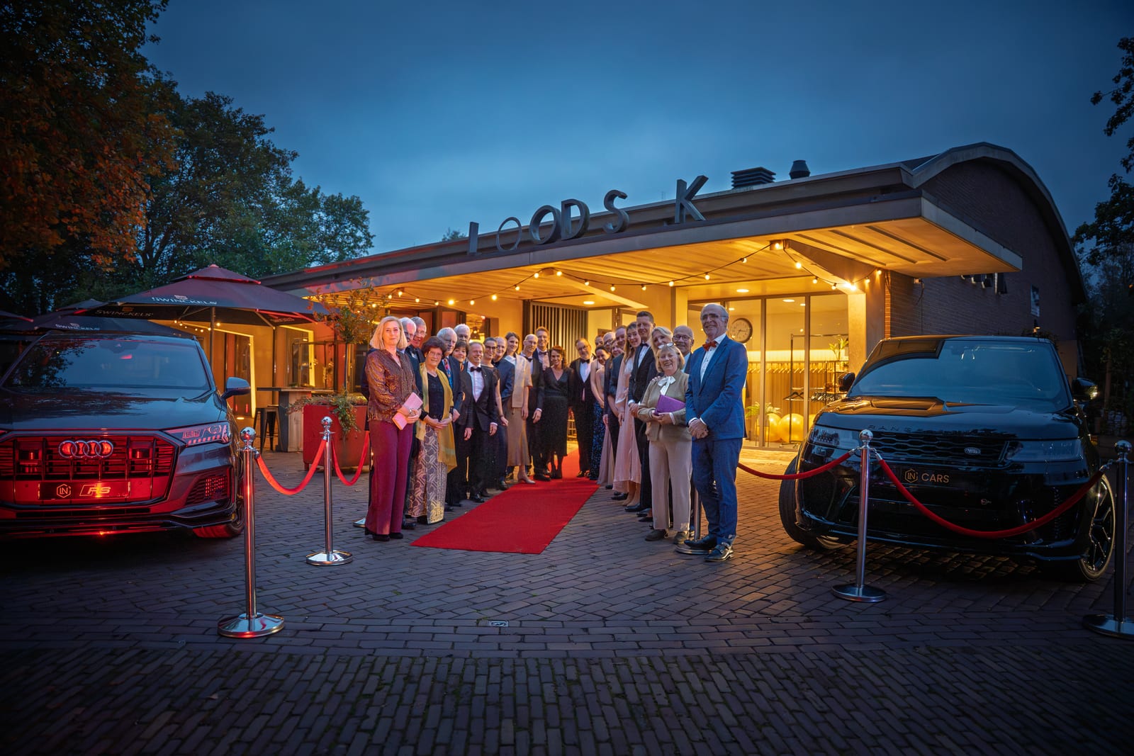 De organisatie van het gala poseert in avondkleding op de rode loper voor de verlichte entree van de loods, tussen twee zwarte auto's onder een diepblauwe avondlucht