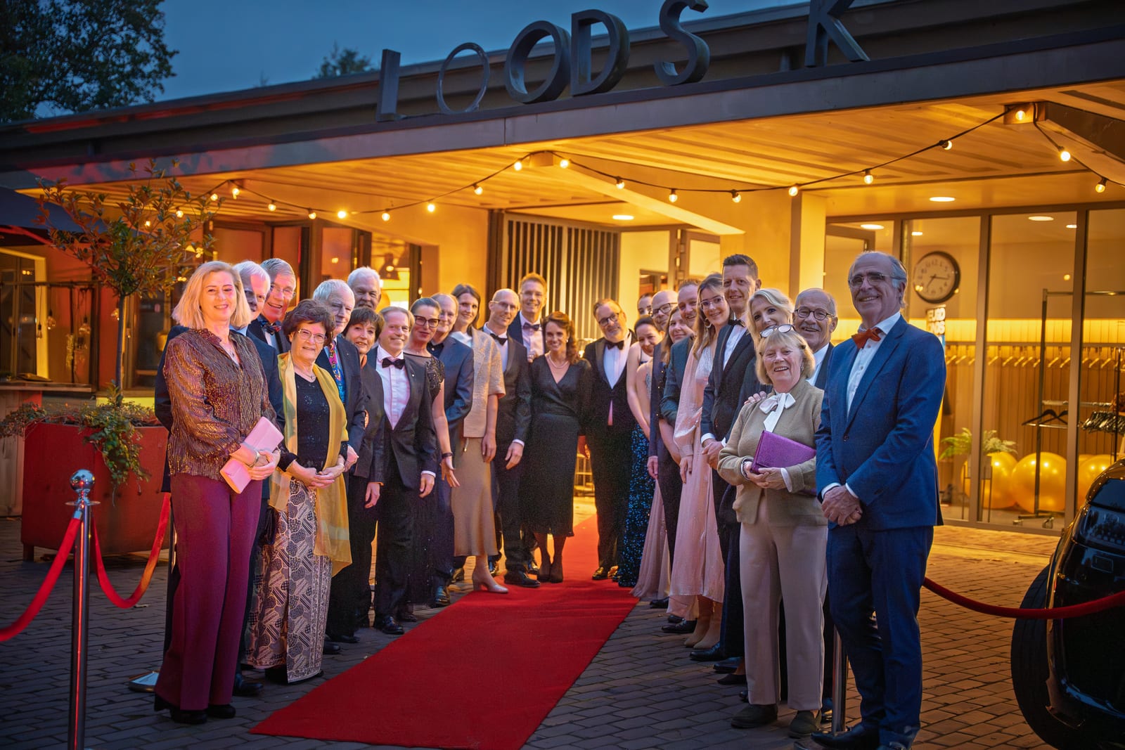 De organisatie staat in een halve cirkel rond de rode loper voor de warm verlichte entree met lampjes en een klok boven de deur