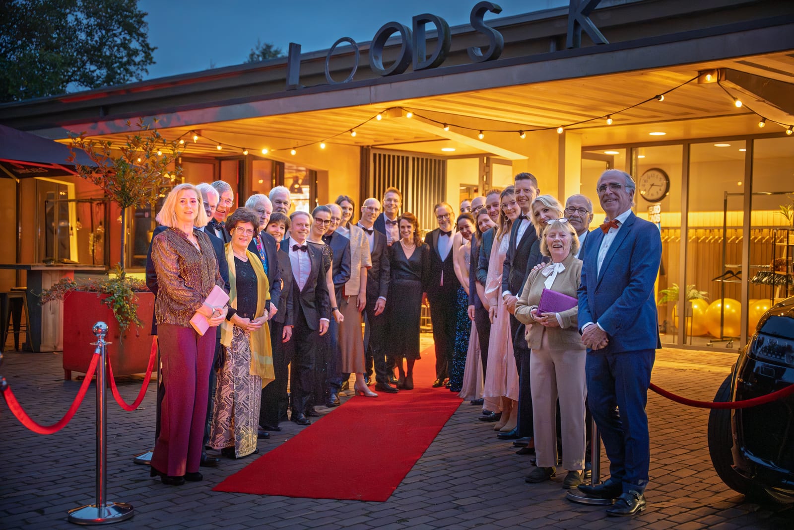 De voltallige organisatie staat opgesteld langs de rode loper onder de lampjes van de entree, met het rode koord op de voorgrond